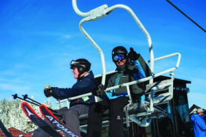 Two people in winter gear sit on a snow-covered ski lift. One gives a thumbs-up, both smiling, with bright blue sky and snowy trees in the background.