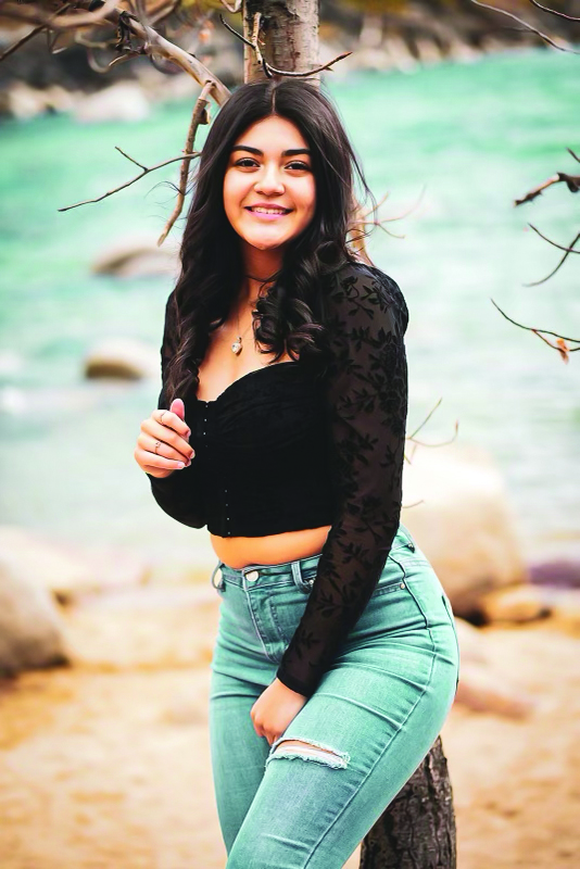 Smiling woman in a black top and jeans stands by a tree, with a blurred background of water and rocks, conveying a relaxed and cheerful mood.